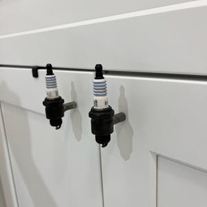 May include: Two black spark plug cabinet pulls mounted on a white cabinet door. The spark plugs have white and blue ceramic insulators and are attached with metal rods. The cabinet door has a simple, clean design.