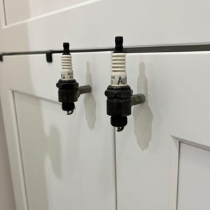 May include: Two spark plug cabinet pulls mounted on a white cabinet door. The spark plugs have a black base, a white ceramic insulator, and a black top. The word "Aut" is visible on one of the spark plugs.