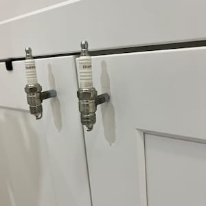 May include: Two spark plug cabinet pulls mounted on a white cabinet door. The spark plugs are silver and white with the word "CHAMP" printed on the side. The cabinet door is white.