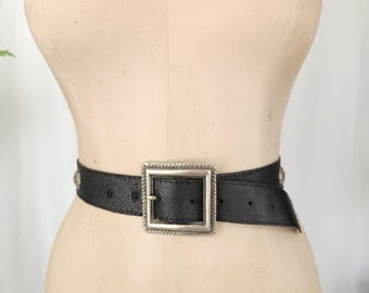 May include: Black belt with a large square silver buckle and silver studs. The belt is worn around a mannequin. The buckle has a decorative border. The belt is made of a textured material.