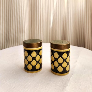 May include: A pair of vintage salt and pepper shakers. The shakers are cylindrical with a black base, adorned with a pattern of yellow teardrop shapes. They have a brass-colored top with small holes for dispensing.