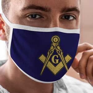 Può includere: Una maschera facciale bianca con sfondo blu e un simbolo massonico dorato. Il simbolo è un compasso e una squadra con una 'G' al centro.