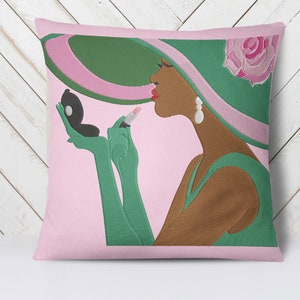 Op de afbeelding: Een roze en groen kussen met een illustratie van een vrouw die een groene hoed met een roze lint draagt en lippenstift aanbrengt. De vrouw draagt een groene jurk en handschoenen.
