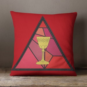 Puede incluir: Cojín rojo con un diseño de triángulo negro que presenta un cáliz dorado en estilo de vidriera.