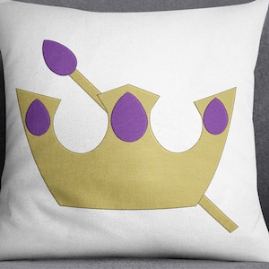 May include: A white cushion featuring a gold crown with purple accents. The crown has three points and a purple sceptre.