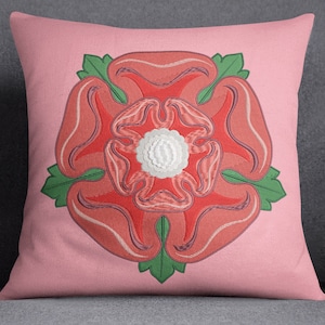Op de afbeelding: Een roze kussen met een geborduurd rood rozenmotief. De roos heeft een wit centrum en groene bladeren.
