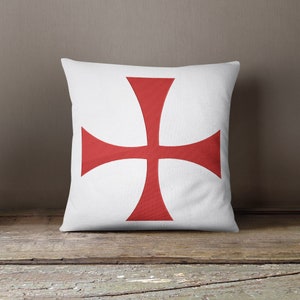 Può includere: Un cuscino bianco con una croce rossa al centro. La croce è un design semplice e simmetrico con bordi arrotondati.