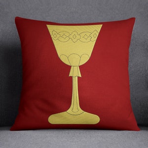 Puede incluir: Una almohada roja con un diseño bordado en oro de una copa. La copa tiene un borde decorativo y un tallo alto.