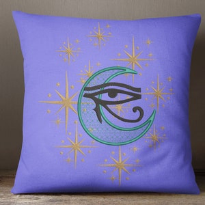 Puede incluir: Una almohada morada con un diseño bordado de una luna creciente con un ojo dentro. La luna es de color verde azulado y el ojo es negro. El fondo está lleno de estrellas doradas.
