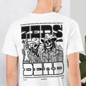 May include: White t-shirt with a black and white graphic print of two skeletons wearing cowboy hats and holding guns. The text "ZEPS DEAD" is printed in large black letters above the skeletons. The text "DC HOOKS" is printed in smaller black letters above the skeletons. The text "VILLA KILLA" is printed in small black letters at the bottom left of the graphic. The text "CASHMERE DESIGNS" is printed in small black letters at the bottom center of the graphic. The text "JUNE 2021" is printed in small black letters at the bottom right of the graphic.