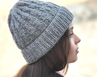Puede incluir: Un gorro de punto gris con un borde acanalado grueso y un patrón de puntadas verticales texturizadas. El gorro está hecho de hilo y presenta un diseño de ala doblada que se ajusta a la cabeza, ofreciendo una cobertura total en tono gris.