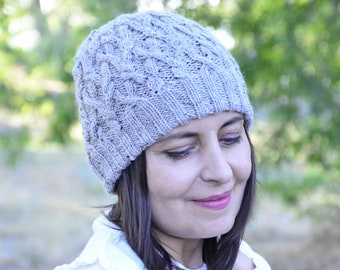 May include: A woman wearing a light brown knitted hat with a cable knit pattern. The hat is pulled down over her forehead and she is looking down.