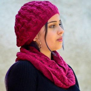 以下が含まれることがあります： ピンクのニットのベレー帽とそれに合わせたスカーフを着用した女性のクローズアップ。ベレー帽はテクスチャのあるパターンで、スカーフはシンプルなニットです。