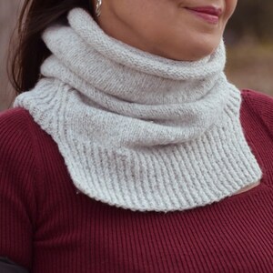 May include: A light gray knitted cowl scarf. The scarf is made of a soft, textured yarn and has a ribbed design at the bottom. The cowl is worn around the neck and is a neutral color.
