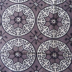 May include: Close-up of a fabric with a repeating pattern of circular designs. Each circle features a central floral motif surrounded by intricate, swirling patterns in white against a muted purple background. The design is reminiscent of a mandala.