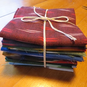 May include: A stack of colorful fabric squares tied with twine. The top fabric is a deep burgundy with red and white stripes. Below are layers of blue, green, and other hues. The bundle is neatly arranged, suggesting a craft or sewing project.