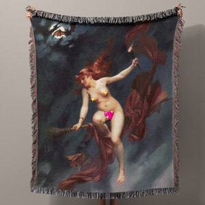 May include: A woven tapestry throw blanket with a dark blue background. The blanket features a painting of a woman flying on a broomstick with a red cape. The woman is nude and has long red hair. The painting is set against a dark blue sky with clouds and a bat flying in the upper left corner.