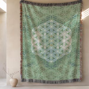 May include: A green throw blanket with a geometric flower of life pattern. The blanket has a fringe border and is displayed on a wall. The design features a central flower of life motif in white and light blue.