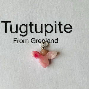 May include: A pendant featuring a raw, pink tugtupite stone shaped like a butterfly with a small, round, peach-colored bead at the top, attached to a silver-toned metal jump ring. The text above reads Tugtupite From Greenland.