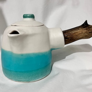 May include: A ceramic teapot with a white and turquoise glaze. The teapot has a wooden handle and a small spout. The lid and the top of the handle are a darker turquoise colour. The teapot is on a white surface.