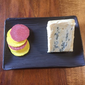 May include: A black rectangular serving platter with a wedge of blue cheese and a stack of red and yellow crackers. The cheese has blue veins, and the crackers are round.