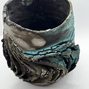 May include: A handcrafted ceramic vase with a textured finish. The vase displays a blend of black, grey, and turquoise hues. The design has a distinctive, organic form with a cracked, weathered look, adding to its unique appeal.