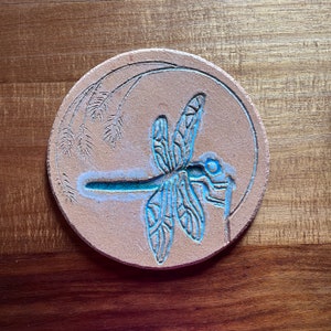 Puede incluir: Un posavasos redondo de cerámica color beige con un diseño detallado de libélula. La libélula está delineada en turquesa, con alas y cuerpo grabados. El posavasos también presenta un diseño de espiga de trigo grabado y un borde turquesa.