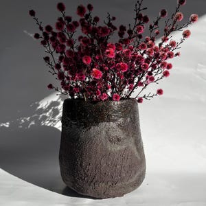 May include: A dark grey, textured ceramic vase with a rough, uneven surface and a darker, smooth rim. The vase is filled with a dense arrangement of thin, branching stems featuring numerous small, vibrant pink, pom-pom-like flower blossoms.