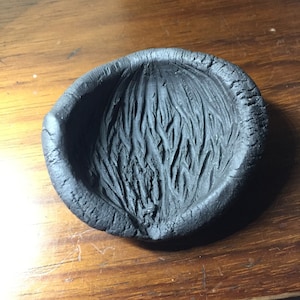May include: A handmade, black ceramic bowl with a textured interior resembling fur or hair. The bowl has a rounded shape with a slightly raised rim, and the surface shows visible imperfections, adding to its rustic charm.