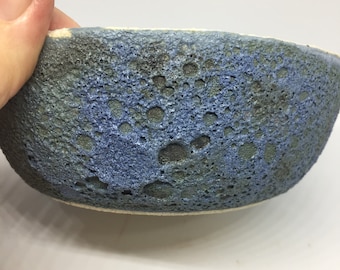 May include: A small ceramic bowl with a textured surface. The bowl has a mix of blue, green, and dark brown colours. The interior is a lighter blue, and the rim is a creamy white.