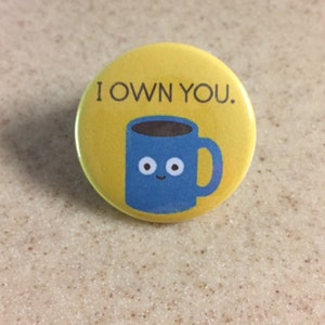 Puede incluir: Un botón amarillo con una taza de café azul con ojos saltones y el texto "I OWN YOU."