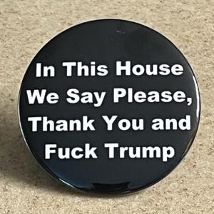 Puede incluir: Un botón negro con texto blanco que dice "In This House We Say Please, Thank You and Fuck Trump."