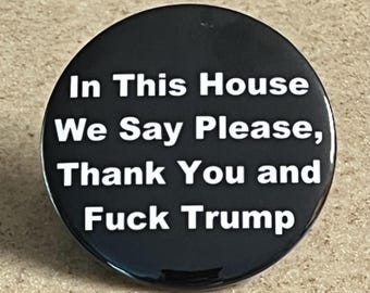 May include: A black button with white text that reads "In This House We Say Please, Thank You and Fuck Trump."