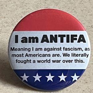 May include: A round button with a red and blue design. The top is red, the bottom is blue with white stars, and the center has the text "I am ANTIFA" with a statement about opposing fascism.