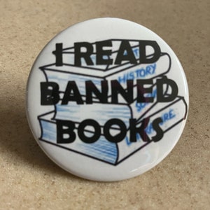 Może przedstawiać: Biała przypinka z czarnym napisem "I READ BANNED BOOKS" nad grafiką trzech książek z niebieskimi liniami. Na książkach widnieją słowa "HISTORY", "SCIENCE" i "LITERATURE".