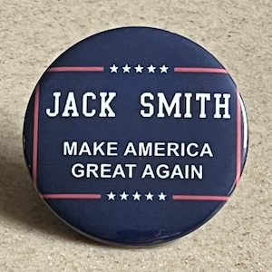 Pode incluir: Um botão redondo azul marinho com texto branco que diz "JACK SMITH" e "MAKE AMERICA GREAT AGAIN". O botão tem uma borda rosa e branca e pequenas estrelas brancas acima e abaixo do texto.