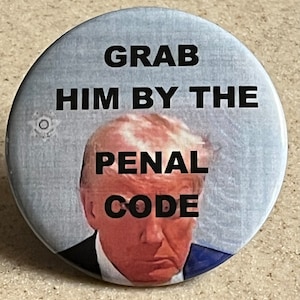 Puede incluir: Un botón redondo con una imagen de Donald Trump y el texto "Grab Him By The Penal Code".