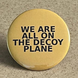 May include: A circular yellow pin-back button featuring the black text WE ARE ALL ON THE DECOY PLANE centred on the face. The button has a smooth, glossy finish and a simple, clean design with bold, sans-serif lettering against the solid yellow background.