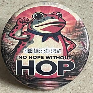May include: Round button with a cartoon frog illustration. The frog is red with black spots, wearing a blue neckerchief, and flexing its arm. Text on the button reads "RIBBIT.RESIST.REPEAT" and "NO HOPE WITHOUT HOP". The background features a sunset scene.