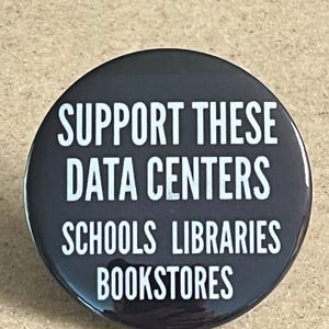 May include: A round, black button with white text. The text reads "SUPPORT THESE DATA CENTERS SCHOOLS LIBRARIES BOOKSTORES". The button is glossy and appears to be made of metal or plastic.