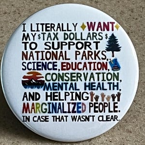 Op de afbeelding: Witte knop met zwarte tekst die luidt: "Ik wil letterlijk mijn belastinggeld gebruiken om nationale parken, wetenschap, onderwijs, natuurbehoud, geestelijke gezondheid en het helpen van gemarginaliseerde mensen te ondersteunen. Voor het geval dat niet duidelijk was."