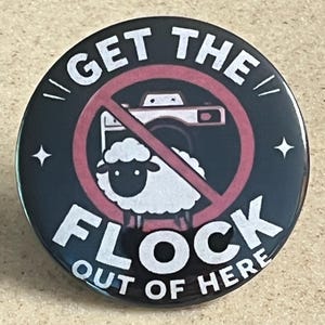 May include: A circular black pin-back button featuring a white sheep graphic centered behind a red prohibition symbol. The text GET THE FLOCK OUT OF HERE is printed in white capital letters around the sheep and a camera icon.