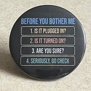 May include: A black button with white text that reads "BEFORE YOU BOTHER ME" and lists four questions: "1. IS IT PLUGGED IN?", "2. IS IT TURNED ON?", "3. ARE YOU SURE?", and "4. SERIOUSLY, GO CHECK".