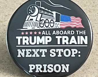 May include: A black and white button with a train and an American flag. The text reads "All Aboard The Trump Train Next Stop: Prison."