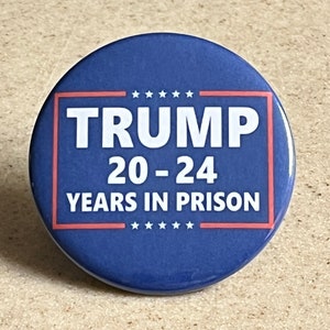 May include: A round, blue button with the word "TRUMP" in white block letters, framed in red. Below, the text reads "20-24 YEARS IN PRISON" in white. Six white stars are above and below the text.