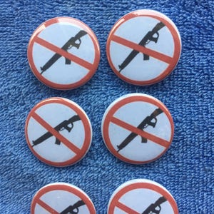 May include: Six white and red round buttons with a black silhouette of a rifle with a red line through it. The buttons are on a blue fuzzy surface.