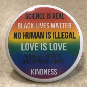 May include: A round button with a rainbow background and white text. The text reads "Science is real, Black Lives Matter, No Human is Illegal, Love is Love, Women's Rights are Human Rights, Kindness."