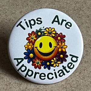 Könnte beinhalten: Ein weißer Button mit dem grünen Schriftzug "Tips Are Appreciated". Das Design zeigt ein gelbes Smiley-Gesicht, umgeben von einem Kranz aus bunten Blumen in Rot, Gelb, Blau und Lila.