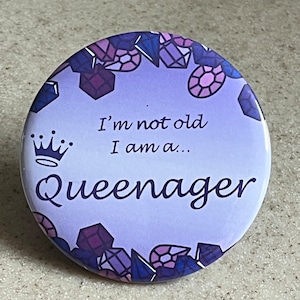 May include: Purple button with a crown and the text "I'm not old I am a... Queenager" surrounded by a border of purple, pink, and blue gemstones.