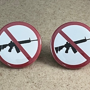 Peut inclure: Deux badges bouton rouge et blanc avec une silhouette noire d'un fusil avec une ligne rouge qui le traverse. Le texte "No Guns" est imprimé sur les badges.